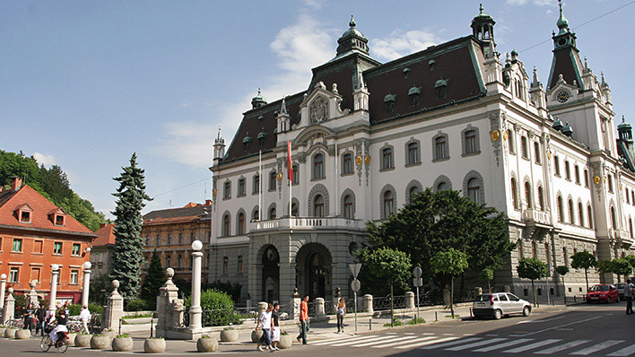 The ornate facade of the University of Ljubljana, featuring intricate architectural designs and adorned with flags, with pedestrians walking in the foreground on a sunny day.