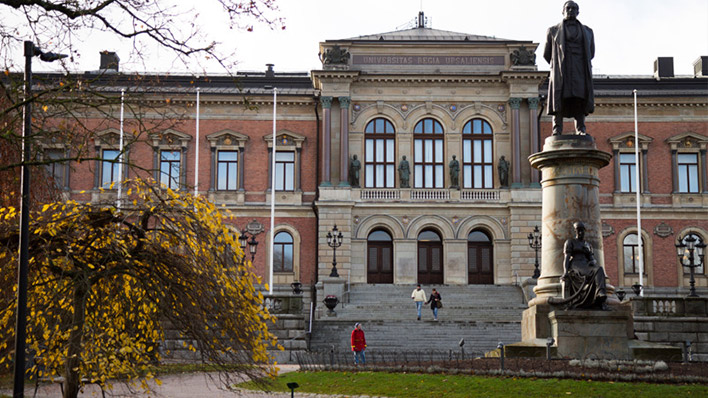 The historic building of Uppsala University, with its classic architecture, is viewed from the park with a statue in the foreground and autumnal trees adding a splash of yellow to the scene.