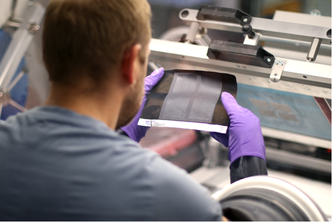 A technician in a lab, viewed from behind, inspecting a flexible, printed battery sheet with a translucent appearance. He is wearing purple gloves, indicating a clean and controlled environment for advanced battery technology research.