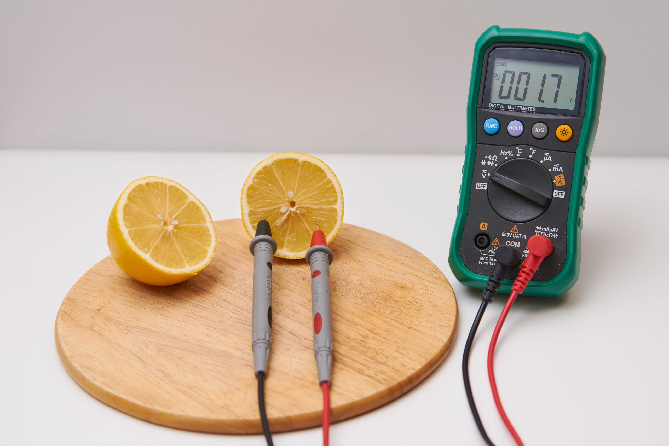 A digital multimeter displays a voltage reading from a lemon battery experiment, with two halves of a lemon connected by probes on a wooden cutting board, demonstrating an educational example of generating electricity from citrus fruit.