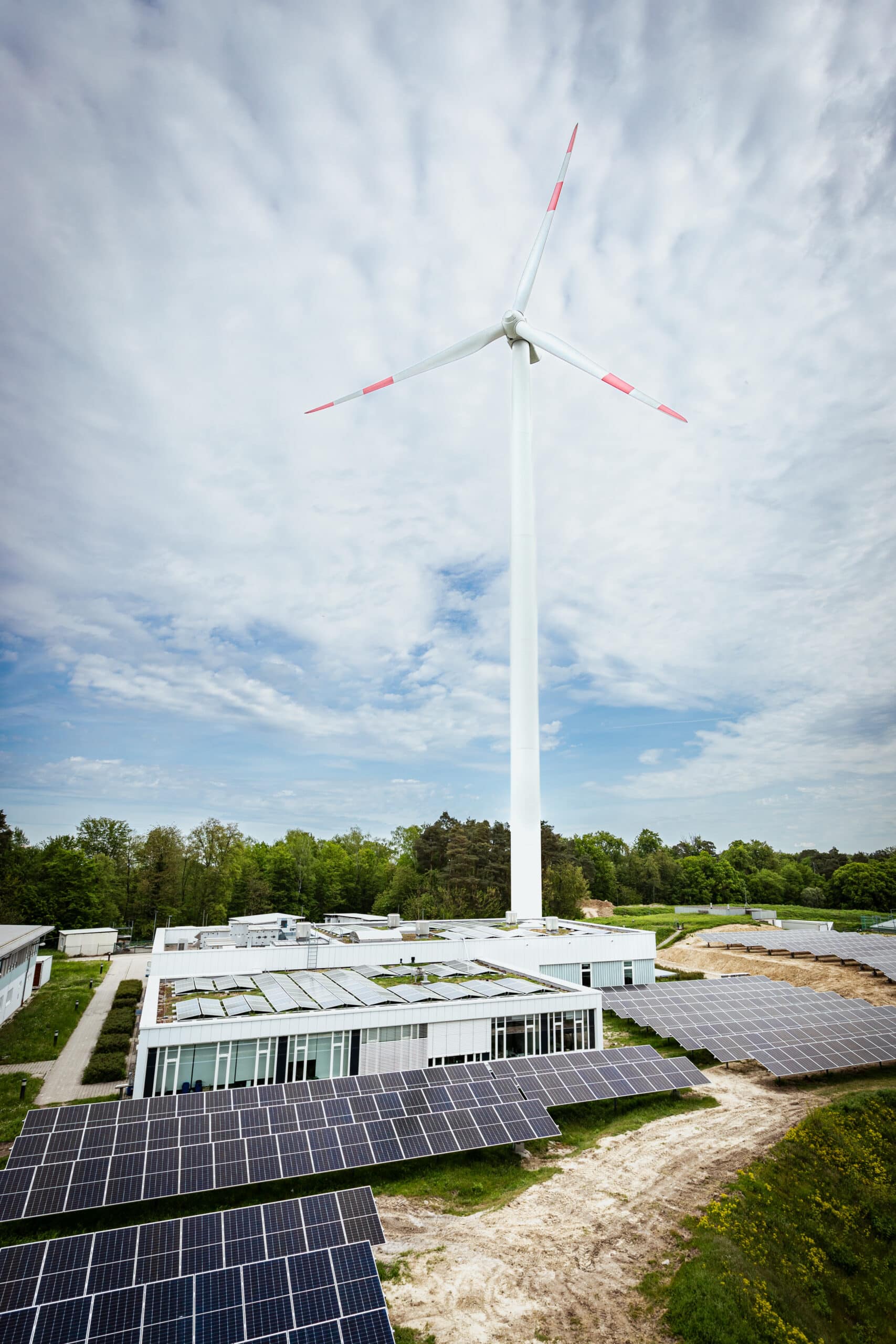 a windmill stands behind a couple of low buildings that hold the redox-flow batteries and there are solarpanels surrounding them.